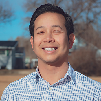 Head and shoulders of John Geyerman, Asian decent, short dark hair, white button-down shirt with light blue grid lines, standing in open field early spring