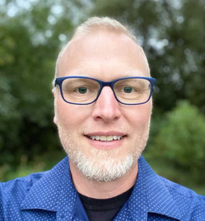 Head and shoulders of Matthew Anderson standing in front of green trees blurred in the background, wearing royal blue collared shirt, dark blue rimmed glasses, with short, light hair and short gray beard