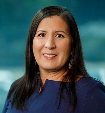 Head and shoulders of Melinda Padilla Lynch with long, straight, dark hair, dark eyes, toothy smile, wearing a navy blue long-sleeve shirt and standing in front of a teal background
