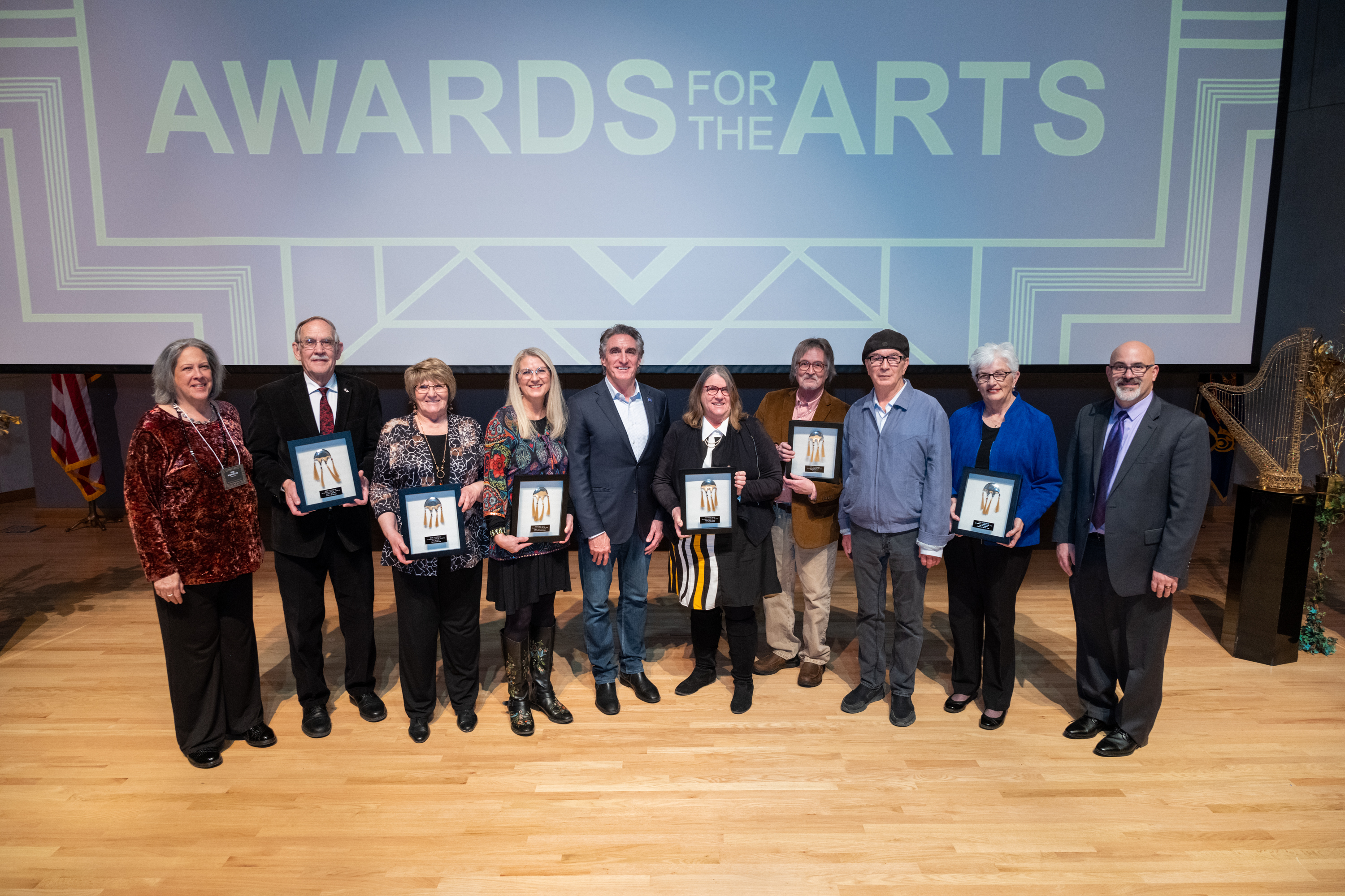 Ten adults standing on a hard wood floor, dressed business casual, holding six large black-framed awards with a large projected screen in the background that reads AWARDS FOR THE ARTS. 