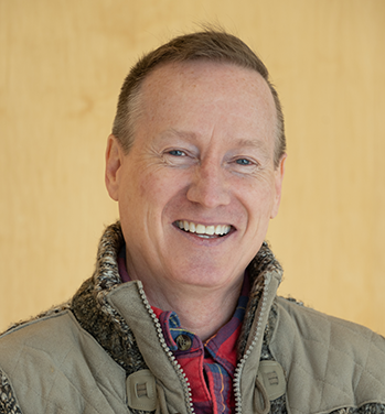 head and shoulders of smiling Troyd Geist wearing tan vest over red flannel shirt, with short brown hair and a cream-colored background 