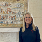 Pieper Bloomquist wearing black long-sleeve shirt, smiling, with glasses and long blonde hair, standing next to her painted tapestry.