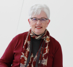 Joan Heckaman wearing deep red sweater, decorative matching scarf, glasses, grey hair and toothy smile with red lipstick