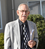 Don Larew standing outside wearing suit with black and white checkered tie, glasses and grey hair