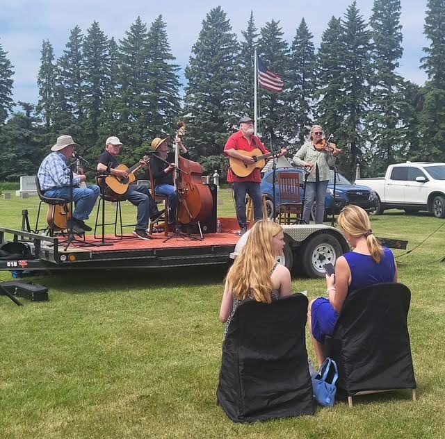 Band of five men plays music, two young women sit in black chairs in front of band