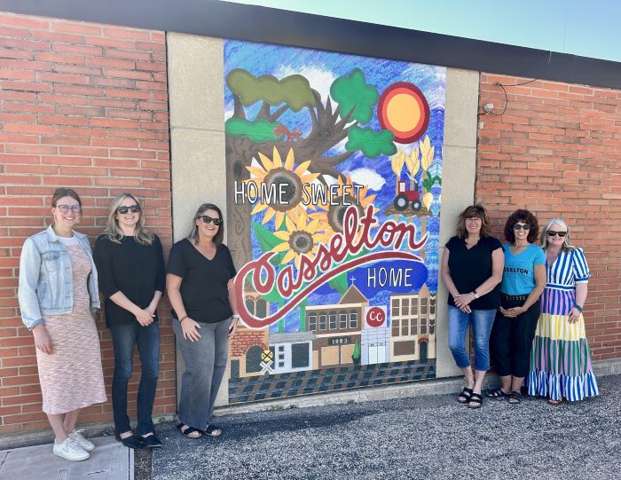 Three women stand on each side of a colorful mural on a brick wall