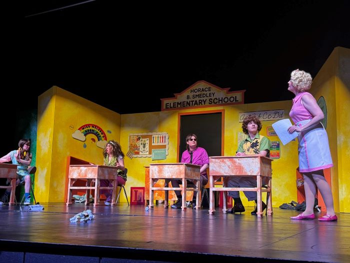 Four people dressed as children sit at desks and one teacher actor on stage with yellow background