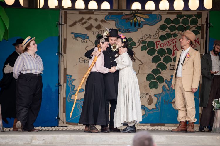 Six people stand on stage, three people hug in center of stage, women in long skirts, men in suits