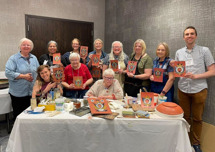 Group of people hold up painted squares, one woman sits at table with painting supplies