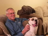 Wayne Beyer wearing glasses, grey hair, smiling, sitting on a chair with white bulldog on his lap