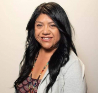 Head and shoulders of Native North Dakota artist Shawna Fricke (Piaute, Taos Pueblo) with long curly black hair, toothy smile, wearing long beaded necklace, patterned shirt and light grey shawl
