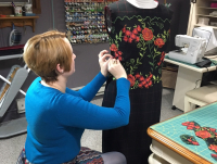 Cynthia McGuire Theil wearing a long-sleeve teal-blue shirt with short red hair, sewing red roses on a black dress
