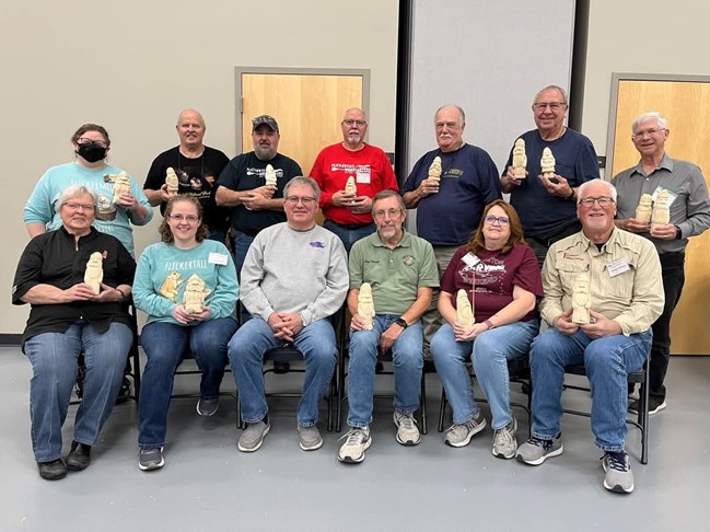 13 people sitting and standing, holding the creations made during the Flickertail Woodcarvers’ 54th Annual Woodcarving Show in Bismarck