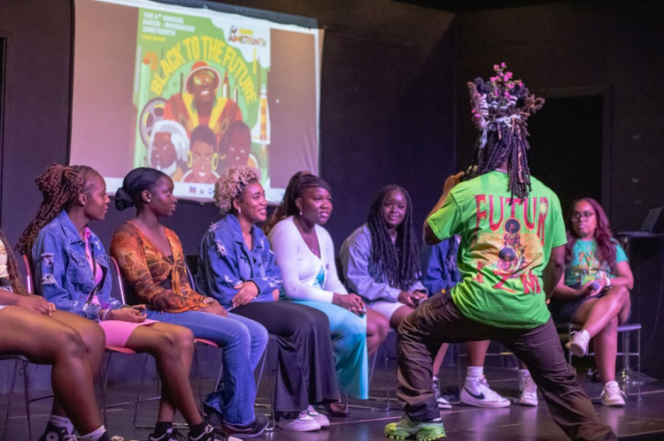 Black to the Future panel discussion during Moorhead Film and Art Festival, celebrating Juneteenth 2023; 8 black women sitting on chairs on a stage with a black man speaking into a microphone in front of them
