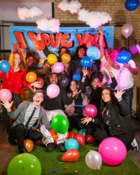 About a dozen teenagers with big smiles and waves, piled together among colorful balloons in front of a poster that says I love you; Fargo Got Talent, Youth Night 2023