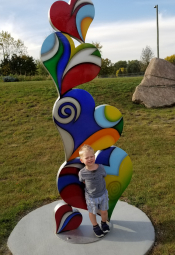Wayne Beyer Grandson Jack standing beside brightly colored 6 foot heart sculpture