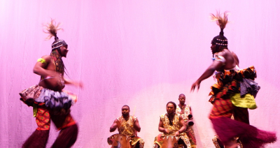 Festival performers in colorful, patterned, ruffled, African clothing, dancing on a stage with Togolese artists drumming in the background
