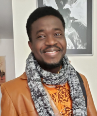 Head and shoulders of Hamzat Koriko with toothy smile, short, black hair, wearing a light brown leather suit jacket, orange patterned undershirt and long black and white scarf wrapped around his neck