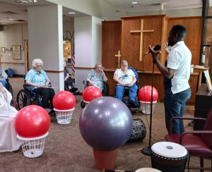 Togolese drumming artist Hamzat Koriko speaking into a microphone to elderly women sitting in wheelchairs with make-shift drums next to them