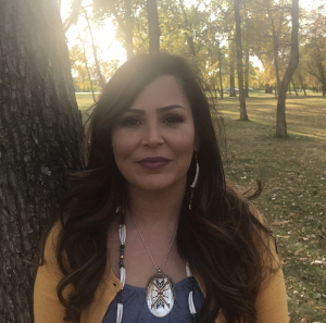 Head and shoulders of Dakota artist Holly Young with long curly brown hair, wearing Native jewelry, standing among tall, leafy trees on a warm sunny day