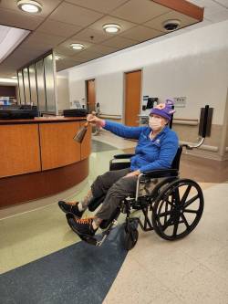 Jane Gaffrey wearing a blue shirt and a pink cartoon beanie sits in a wheelchair in a hospital setting. She is holding a bell up in the air as if ringing it, with a masked smile.