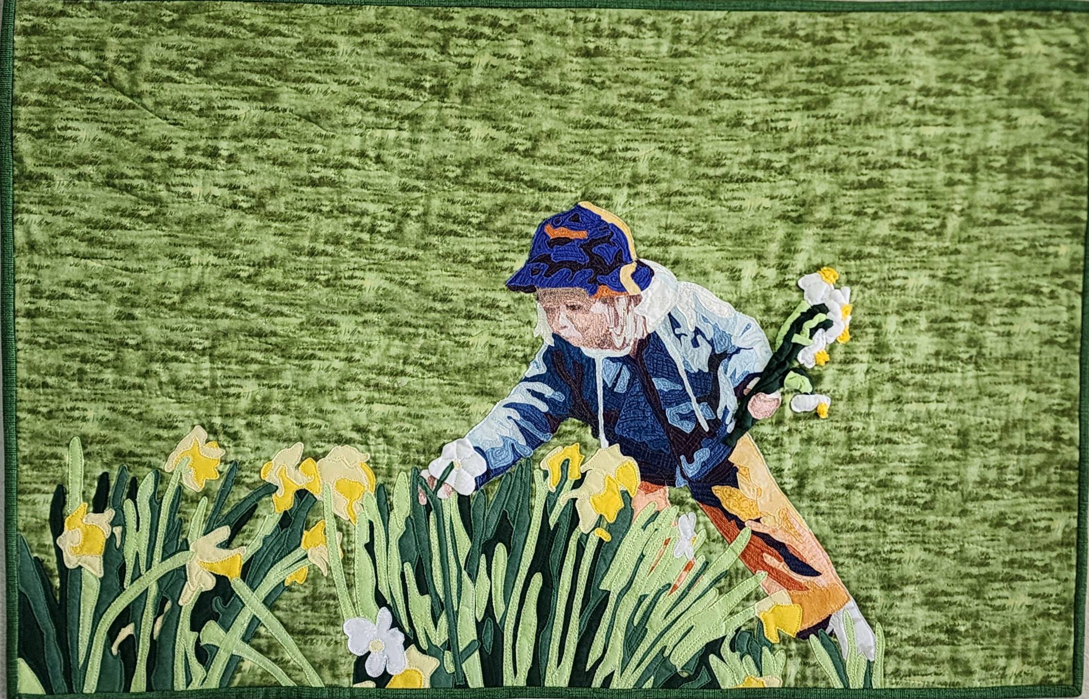Artwork titled Nanna's Daffodils: A vibrant quilt art piece depicting a person in a blue and orange hat and jacket reaching into a field of yellow and white daffodils. The background is a textured, light green fabric.