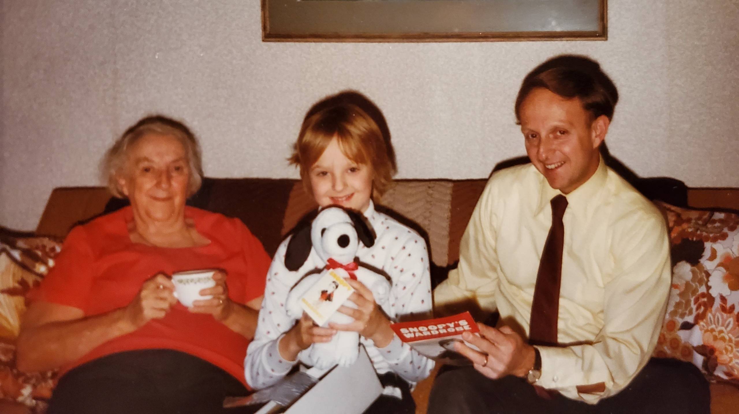 Jane's grandmother (left), Jane as a young girl (center), and Jane's dad on the right, all sitting on a couch. The woman on the left, with short white hair, is wearing a red shirt and holding a white teacup and saucer. The young girl in the middle, with a white patterned shirt and a red bow in her hair, is holding a plush Snoopy doll. The man on the right, wearing a light yellow shirt and a dark tie, is holding a small, red book that says "SNOOPY'S WARDROBE" on the cover. All three are looking at the camera and smiling.