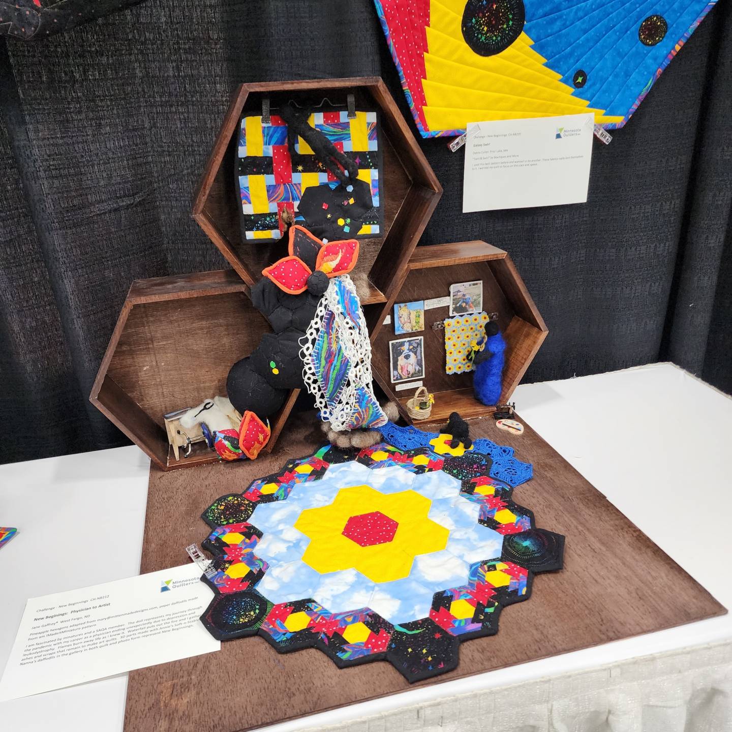 An elaborate quilt display features several pieces of fabric art arranged on a table. A hexagonal quilted piece on the table shows a central yellow and red design, surrounded by a blue and white sky pattern, with a border of black hexagons filled with vibrant colors and stars. Above this, three stacked hexagonal wooden shelves hold smaller quilted items, including a piece with a black and white checkerboard pattern, and another with bright, abstract shapes. There are also small objects like a small toy horse, photos, and a blue felted object. A white paper with text is visible on the table in the lower-left corner.