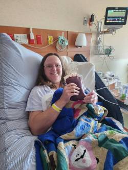 Jane Gaffrey, with long hair and glasses, smiling in a hospital bed, holding up a bag of blood/stem cells. She is wrapped in a quilt and has a hospital bracelet on her wrist.