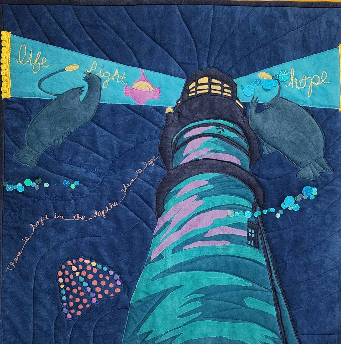 By artist Jane Gaffrey: A quilted art piece depicts a lighthouse against a dark blue, textured background. The lighthouse is decorated with a swirling pattern of teal, purple, and green fabric. Two stylized, dark grey marine animals, possibly manatees or seals, swim on either side of the lighthouse. Above the animals, a blue ribbon-like banner stretches across the top of the image with the words "life," "light," and "hope" embroidered on it in yellow script. The lighthouse shines a yellow light from its top. The phrase "there is hope in the depths there is hope" is embroidered in white script that curves around the left side of the lighthouse. There are also small, colorful fabric embellishments and iridescent sequins scattered around the piece.