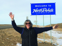 Waste-up photo of Kim Konikow, wearing sunglasses and a black winter jacket, waving hello and posing in front of the Welcome to North Dakota sign on a cold January day with blue skies