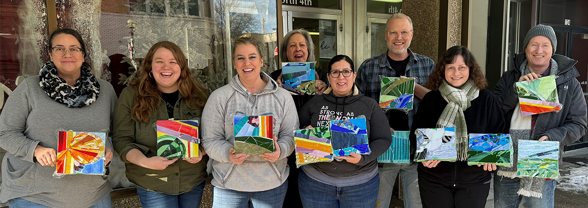 Staff of North Dakota Council on the Arts, eight people standing outside on a cool spring day, smiling brightly and holding artworks they created during a staff retreat