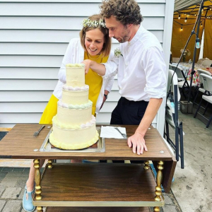 Mollie Douthit and husband Arvid Hägg standing next to and looking own at their tiered yellow wedding cake