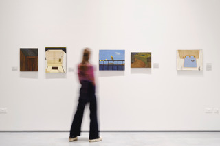 Full-length image of a blurred woman wearing black pants and a long-sleeve dark-pink shirt, standing in between a variety of small rectangular artworks on a white wall