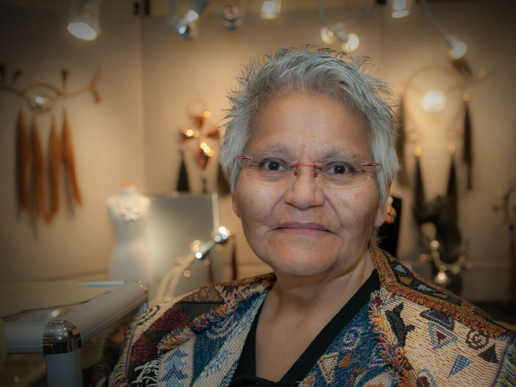 Head and shoulders of Nelda Schrupp with short grey hair, wearing glasses, a shirt with many geometric shapes and colors, and her artistic Native jewelry hanging on the walls behind her