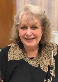 Head and shoulders of smiling Ilonna Pederson with shoulder-length curly blonde hair, aged light skin, black shirt with applique around the neck, holding an oboe