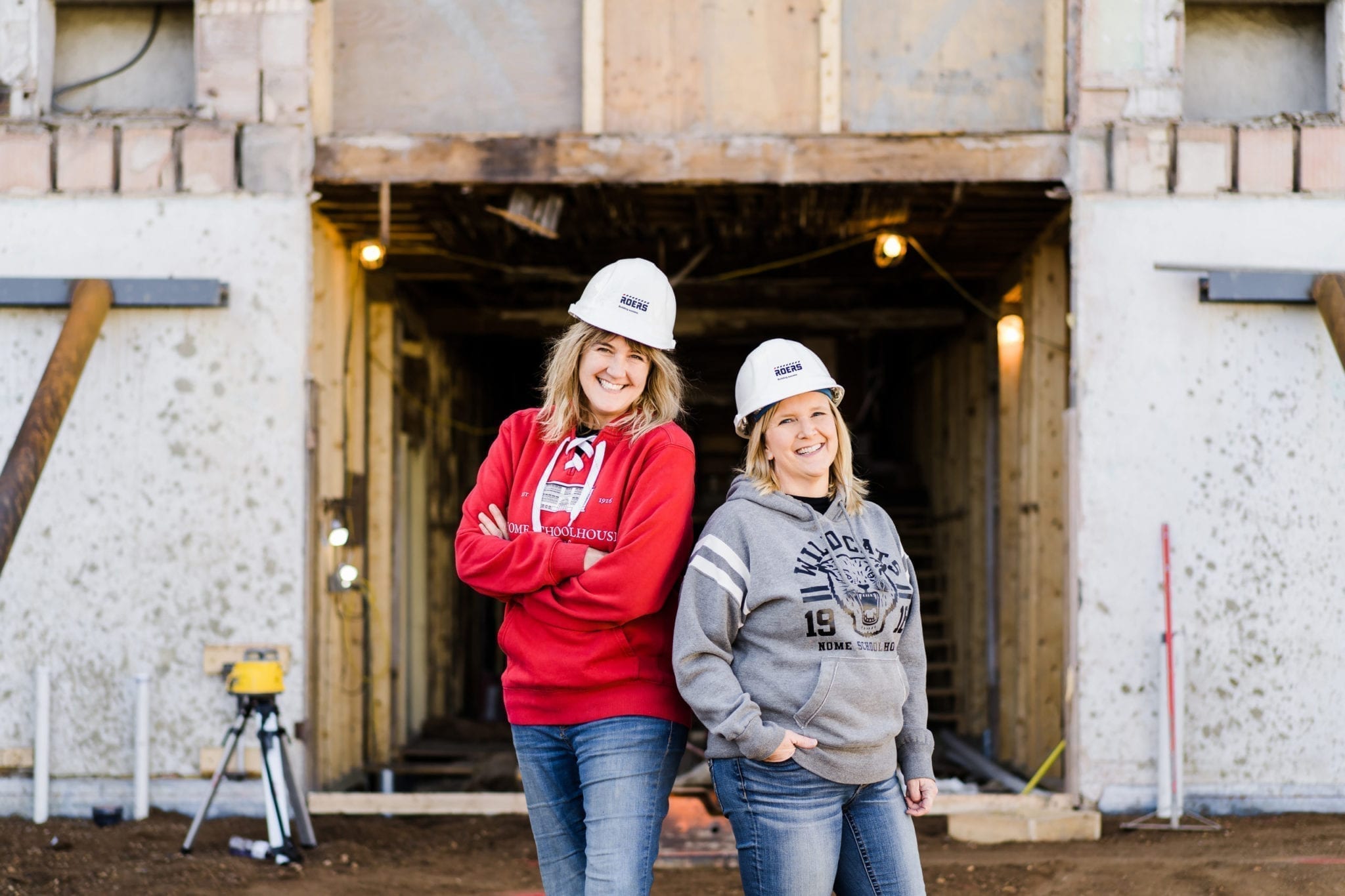 Chris Armbrust and Teresa Perleberg wearing hard hats and Nome Schoolhouse clothing, standing in front of the building when it was still under construction