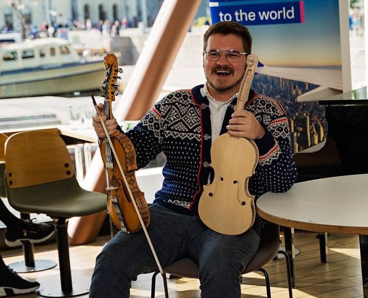Smiling broadly - Gus Holley, of Moorhead, MN, holding two special commemorative fiddles, wearing a Norwegian sweater, sitting on the inner deck of a cruise ship in Oslo, Norway
