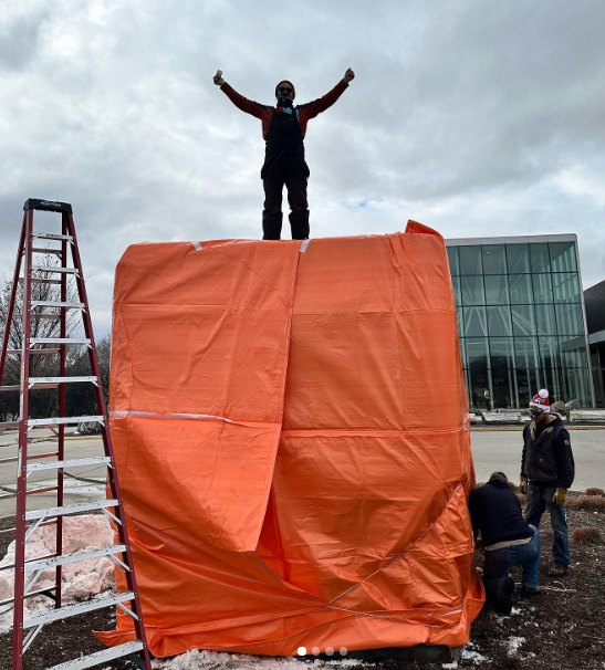 10x10 framed box with snow inside, completed covered by plastic tarps with a man standing on top with his arms outstretched