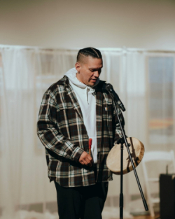 Teaching artist Stuart James standing in front of a microphone and playing a Native American drum