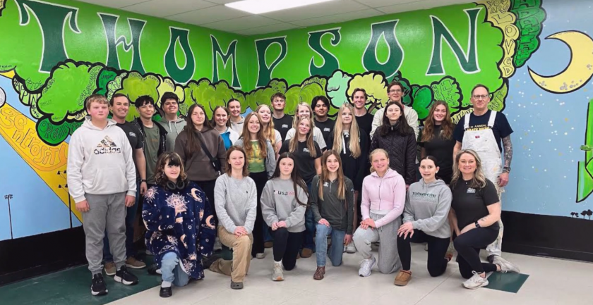 25 students and their teaching artist at Thompson Public school, standing in front of Colorfully painted mural of a huge tree in the center with a sun and rays on the left and a half moon on the right - on two walls in the cafeteria at Thompson Public School; "THOMPSON" in all caps at the top of the tree, inside the leaves; with inspirational words written in the rays, including: determination, faith, adaptable, community, teamwork, grit, collaboration, character, kindness, intelligence, openness, empathy, respect, unity, patience and compassion