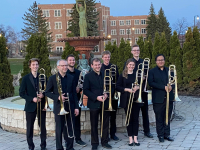 7 men and 1 women in all black, holding trombones, standing on UND Campus
