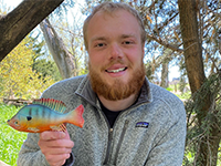 Head and shoulders smiling Walker Bruns with full beard standing among trees and tree trunks with a canopy of leaves holding colorful fish decoy