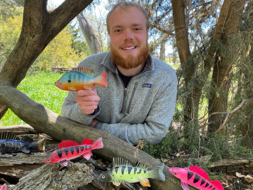 Walker Bruns standing among trees and tree trunks with a canopy of leaves holding colorful fish decoy