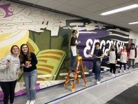 Students at Watford City High School are painting a 20 foot by 10 foot mural (by number) that contains abstract images, including a person reading a book and the word ENGLISH in bright white on top of a dark background containing quotes from famous authors