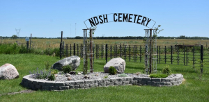 North Dakota State Hospital Cemetery showing green grass and a manicured, decorative area with bricks, rocks and a metal sign that arches over the top saying NDSH Cemetery