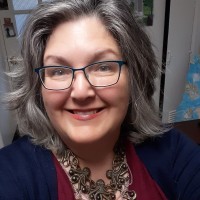 Brightly smiling playwright Kathleen Coudle-King, with shoulder-length dark grey hair, dark clothing, a thickly-designed bronze necklace and thin, dark-rimmed glasses
