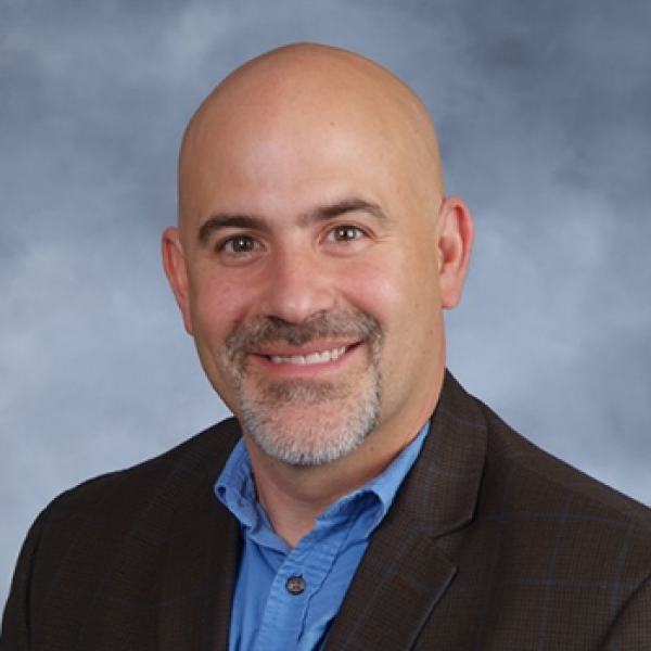 Head and shoulders of Shawn Oban, bald, pale skin, goatee, dark suit jacket with blue button-up shirt on blue cloudy background