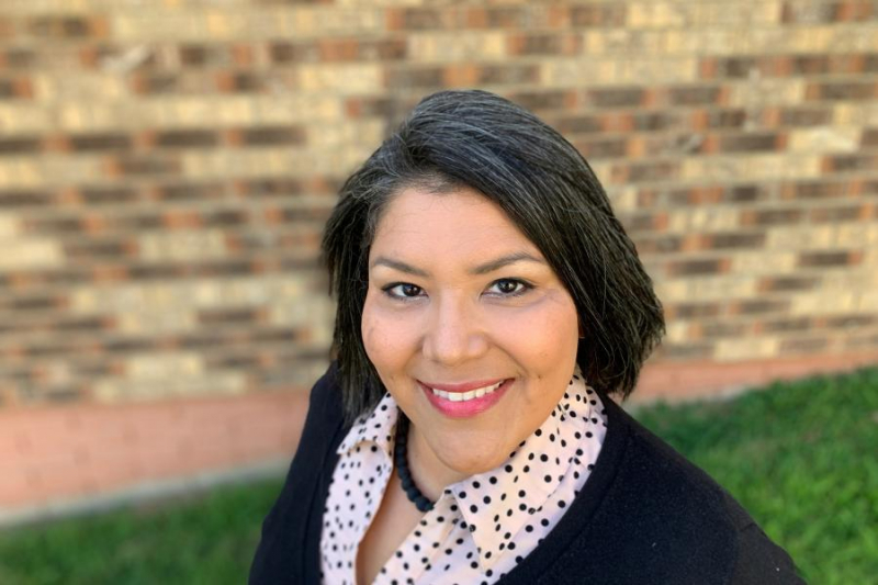 head and shoulders of teaching artist Shelly Sayler with shoulder-length straight, dark hair, dark eyes, a black shirt with a poka-dotted color, thick black necklace, and bright-red lips on a pretty smile