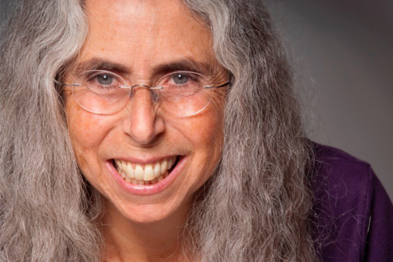 Close-up headshot of Beth Olshansky with big smile, clear glassses and long gray curly hair